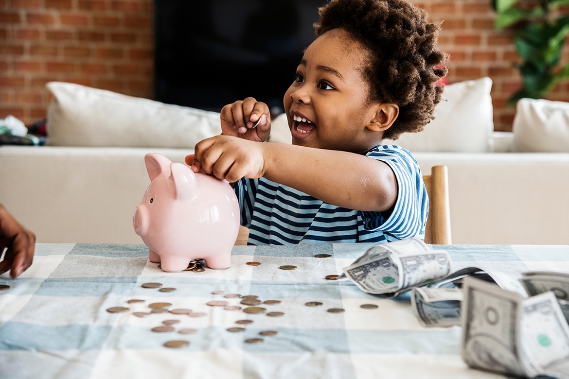 Child placing coins in piggy bank, smiling at table covered with coins and cash, demonstrating budgeting tips.