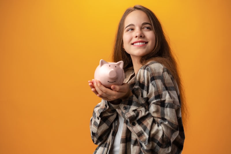 Teen girl in plaid shirt holds a pink piggy bank, smiling against a yellow background, embodying savings tips for beginners.