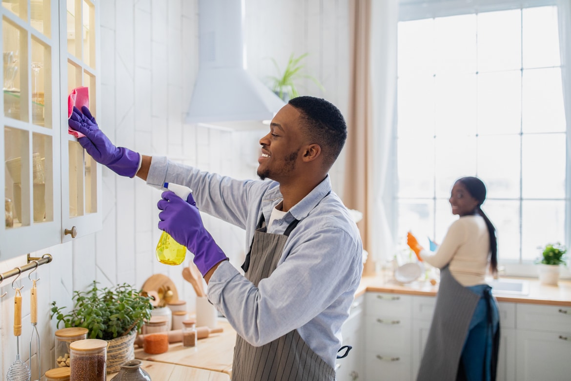Man wearing gloves and apron cleans kitchen cabinet with spray, symbolizing spring cleaning and refreshing finances.