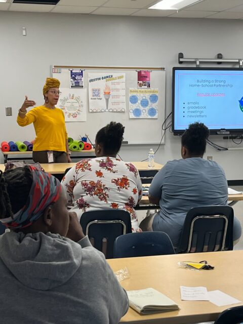 woman in a yellow shirt stands in front of a class speaking