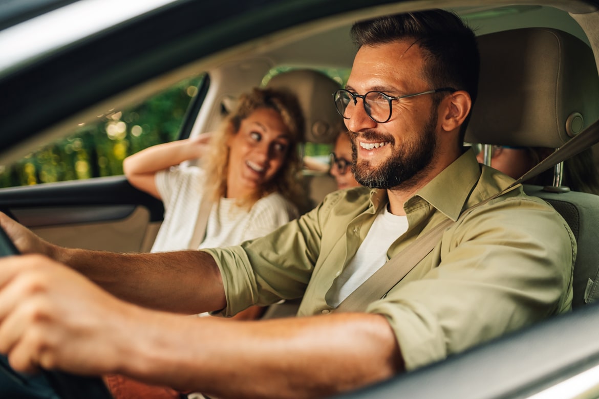 Smiling man driving with family, enjoying a summer car ride, illustrating new or used car-buying guide.