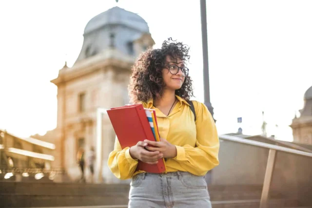 Student holding folders, smiling outside a university building, representing Greenville Federal Credit Union's flexible student loans.