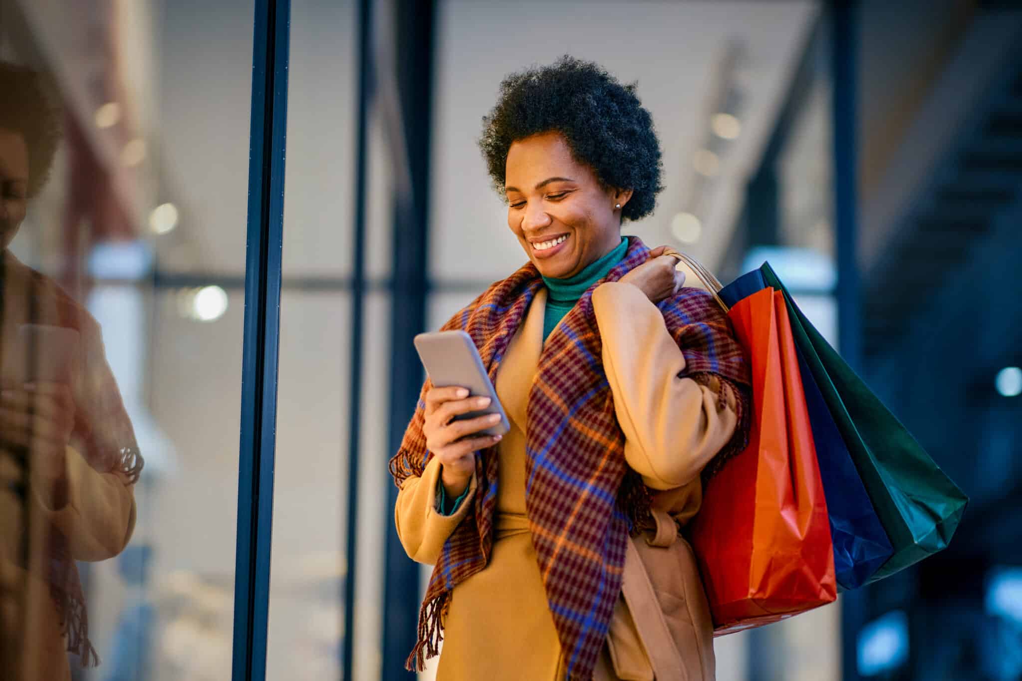 Smiling woman in a scarf checks phone while holding colorful shopping bags, managing cash flow during holiday shopping.