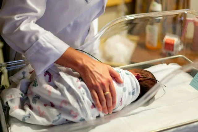 Newborn in hospital bassinet with hand providing gentle support, highlighting life events and local care.