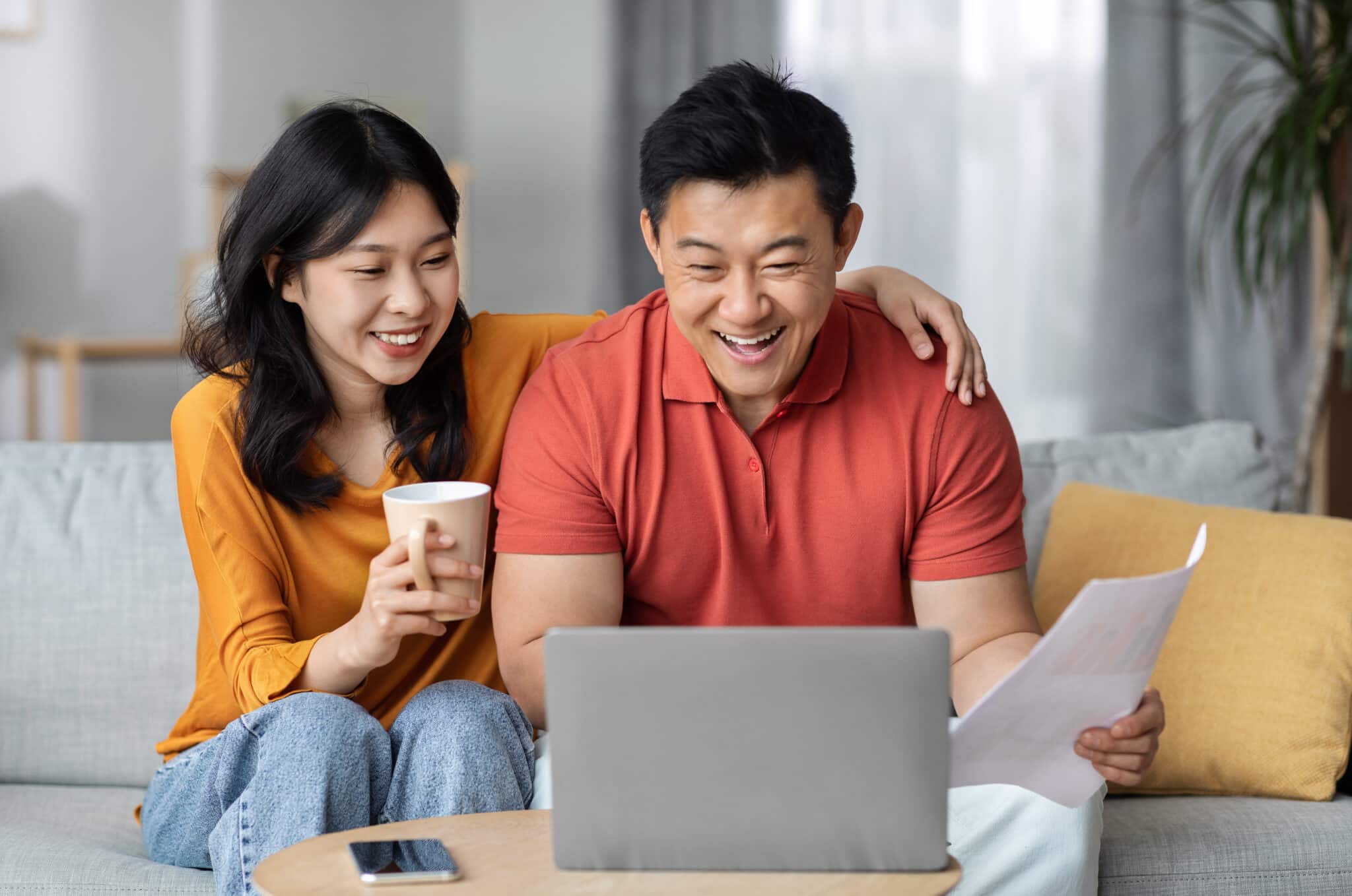 Asian couple managing finances online, sitting on a couch with a laptop and papers, smiling and drinking tea.