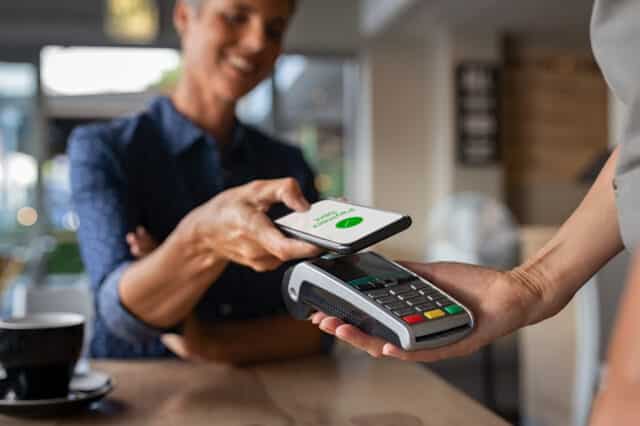 Mature woman uses smartphone for mobile payment via NFC at a coffee shop, illustrating Kasasa Cash Checking benefits.