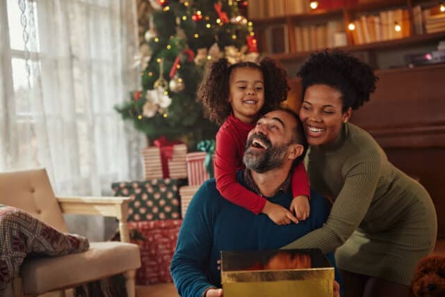 Joyful family with wrapped gifts by a Christmas tree, symbolizing holiday savings for vacations.