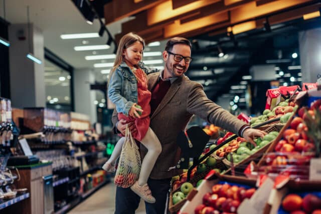 Smiling young father carrying his preschool daughter and they choosing apples together in supermarket