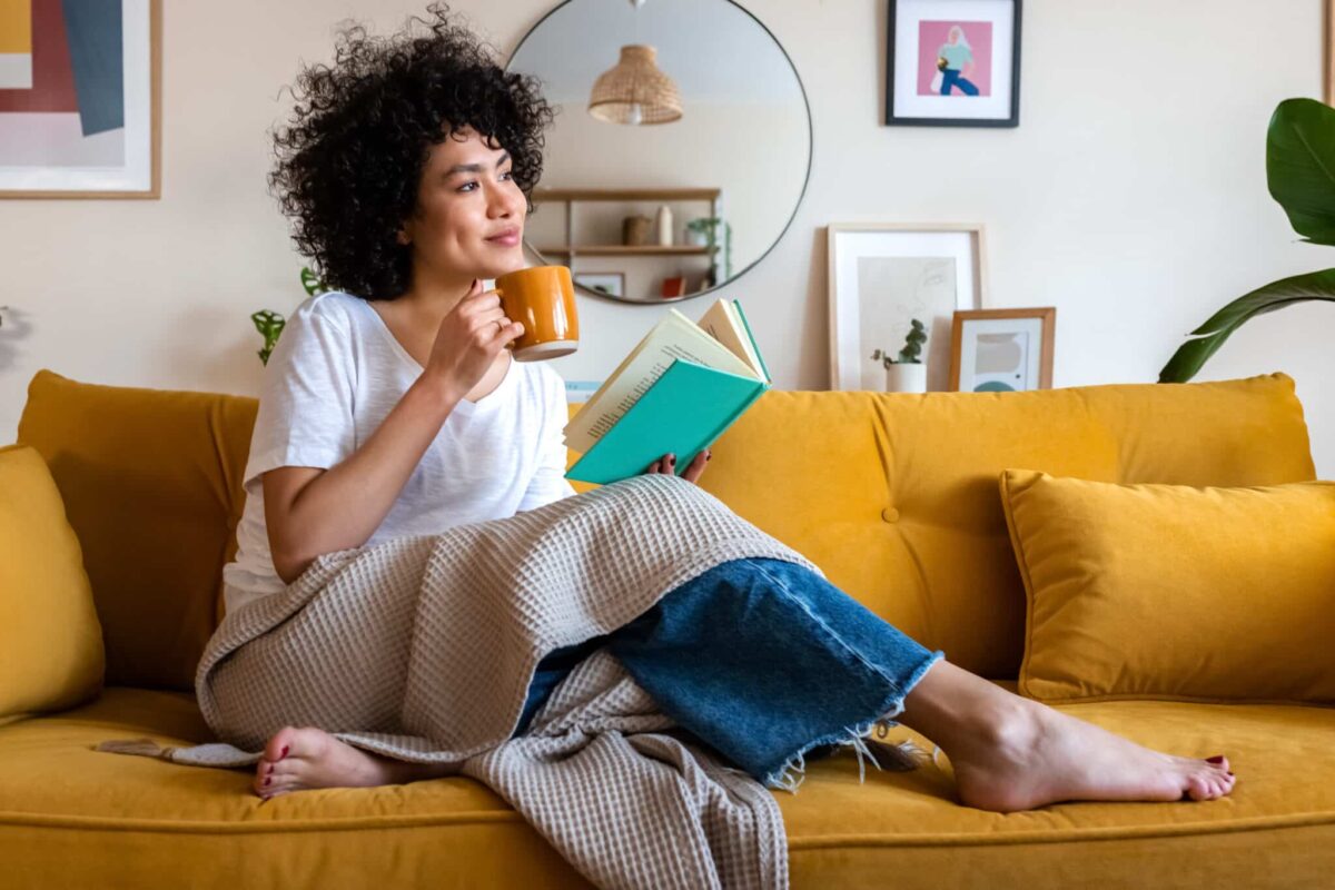 Relaxed woman on couch reading, sipping coffee. Consider skipping your next loan payment; our program fee is $30.