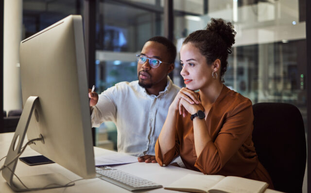 Employees discussing advanced payroll solutions on a computer in a dark office, streamlining HR tasks efficiently.
