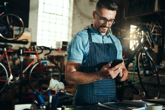 Mechanic in apron uses phone in bicycle shop, exploring tailored commercial benefits for small businesses.