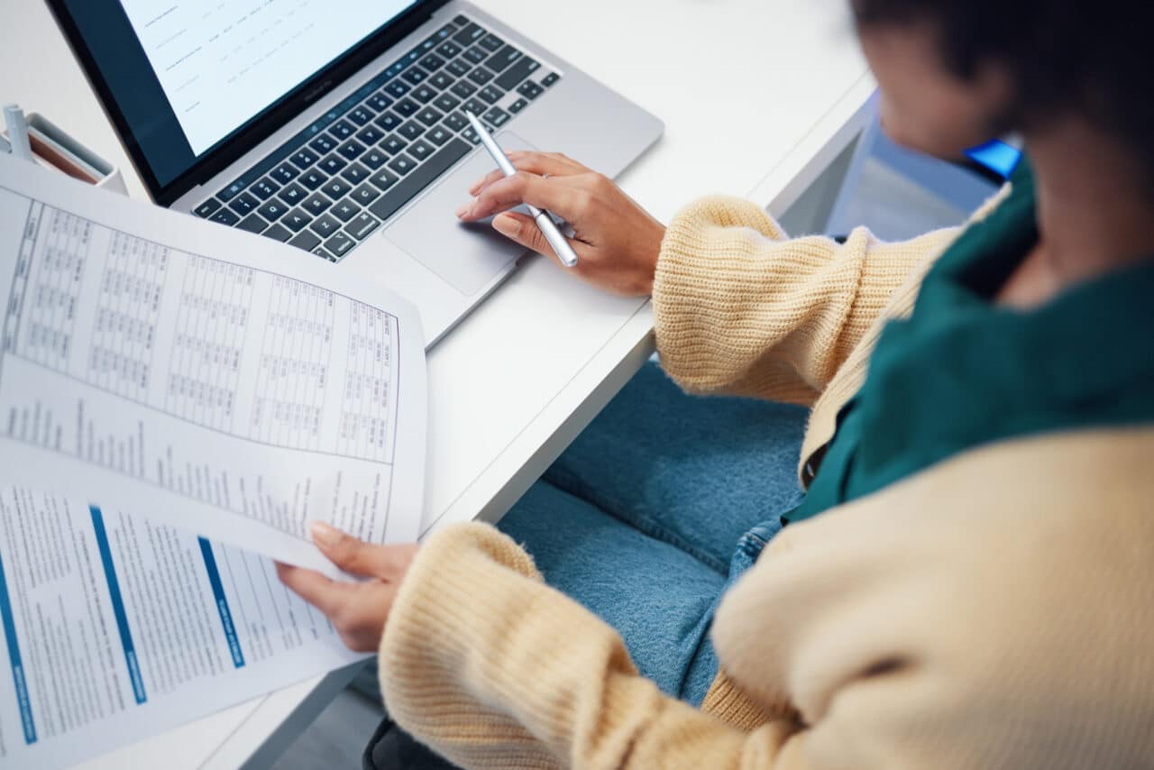 Woman in beige sweater reviewing financial documents and using a laptop for business efficiency in payables solutions.
