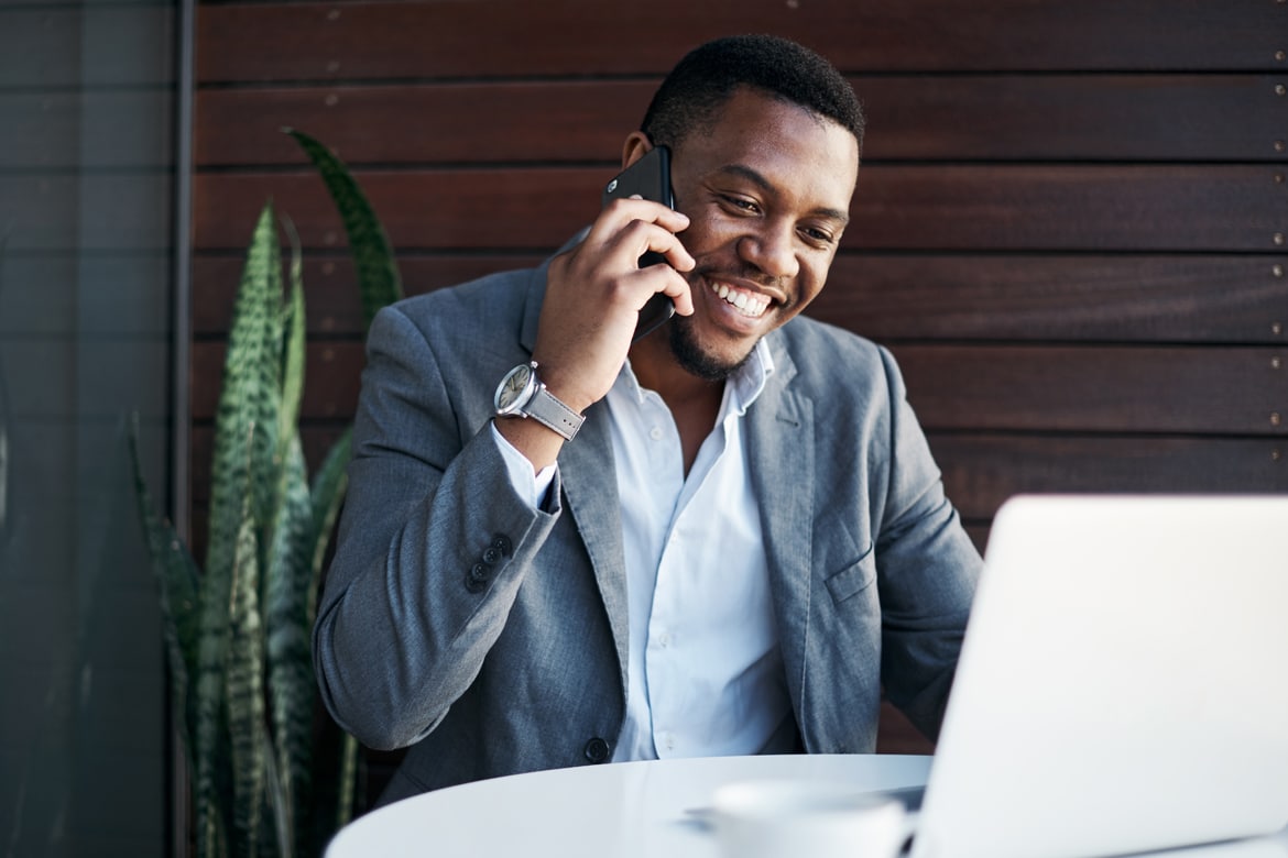 Smiling man in a gray suit talks on a phone while using a laptop, possibly discussing the One Big Beautiful Bill Act's tax effects.