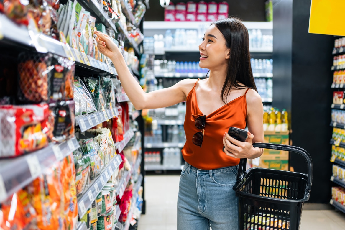 Smiling woman in orange top shops for snacks, illustrating the fine line between savings and overspending.